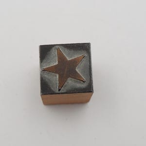 May include: A wooden block stamp with a bronze star design. The star is centered on a gray background, and the stamp has a rectangular shape. The stamp is likely used for crafts or decorative purposes.