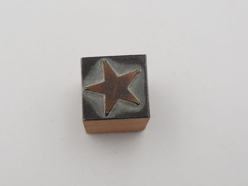 May include: A wooden block stamp with a bronze star design. The star is centered on a gray background, and the stamp has a rectangular shape. The stamp is likely used for crafts or decorative purposes.