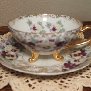 May include: A white teacup and saucer with a gold rim and floral design. The cup has three gold feet and the saucer has a floral pattern with purple and yellow pansies.