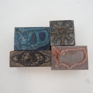 May include: Four vintage woodblock printing stamps. The top stamp is a compass with the words "North, South, East, West" around the compass. The second stamp is a blue and green outline of the United States. The third stamp is a brown woodblock with a globe and the words "Everest, Inc." The bottom stamp is a brown outline of the United States.