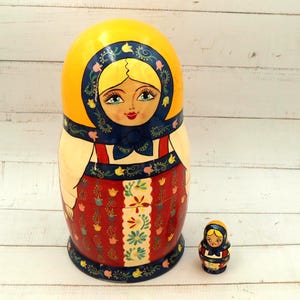 Op de afbeelding: Een grote, handgeschilderde Russische nestpop, of Matroesjka-pop, met een kleinere pop ernaast. De poppen hebben een gele hoofddoek, blauwe accenten en een rode jurk met bloemenpatroon. De grotere pop is ongeveer 20 cm hoog.