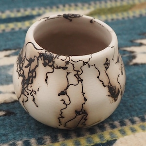 May include: A small, round, off-white ceramic bowl with a distinctive black lightning pattern. The bowl has a wide opening and a smooth, rounded shape. The interior is a light brown colour. The bowl is likely handmade and suitable for decorative or functional use.