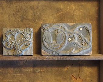 Può includere: Due blocchi di stampa in legno vintage con inserti in metallo. Un blocco presenta un disegno di tre campane con uno stelo. L'altro blocco presenta un disegno di un motivo floreale stilizzato.