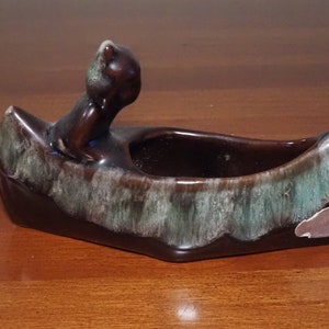 May include: A brown ceramic planter shaped like a canoe with a small brown bear figurine sitting in the front. The planter has a green and white glaze.