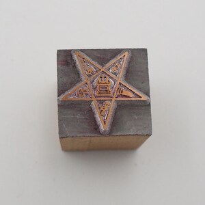 May include: A vintage wood and metal stamp featuring a five-pointed star design. The star has an intricate pattern with copper-colored details. The stamp is rectangular, with the star design centered on the top surface.