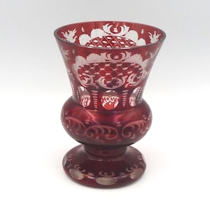 May include: A ruby red glass vase with intricate white etched floral and geometric patterns. The vase has a flared top, a bulbous midsection, and a circular base. The etched designs create a striking contrast against the red glass.