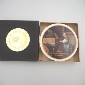 May include: A black box with a gold emblem and a round decorative plate. The plate features a painting of a woman playing a musical instrument, with the text "Detail of LADY SEATED AT A VIRGINAL By VERMEER".