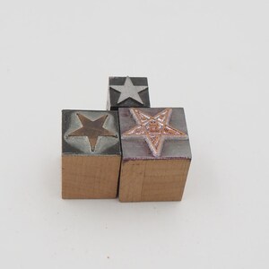 May include: Three wooden block stamps with star designs. One stamp has a silver star, another has a bronze star, and the third has a five-pointed star with a detailed design. The stamps are on a white background.