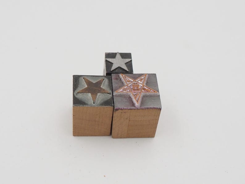 May include: Three wooden block stamps with star designs. One stamp has a silver star, another has a bronze star, and the third has a five-pointed star with a detailed design. The stamps are on a white background.