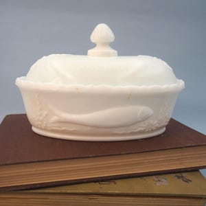 May include: An oval white milk glass dish with a matching lid featuring a finial handle. The exterior surface is embossed with a fish motif and floral details. The lid has the word SARDINES embossed in the centre of the glass surface.