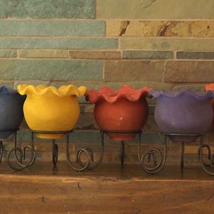 May include: Five colorful ceramic flower pots in blue, yellow, red, purple, and orange, each with a ruffled rim. They are mounted on decorative black metal stands with scrollwork and heart-shaped details. The background is a textured stone wall.