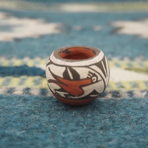 Peut inclure: Petit pot rond en céramique peinte à la main, intérieur et base rouge-orangé. L'extérieur présente un fond blanc avec des motifs géométriques noirs et blancs et un motif d'oiseau rouge. Le pot est probablement un objet décoratif ou une perle.