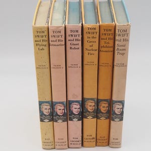 May include: A collection of vintage Tom Swift and His Adventures books. The spines are in various colors, including tan, peach, and orange. Each book features a portrait of a young man and the title of the book.
