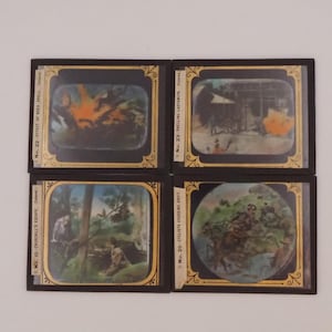 May include: Four antique glass slides with black frames and gold trim. Each slide features a different scene, including explosions and figures in a landscape. The slides are labeled with text, including "EFFECT OF HIGH SHELL" and "CYCLISTS CROSSING DRIFT."