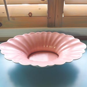 May include: A pink, scalloped-edge ceramic bowl. The bowl has a wide, shallow design with a smooth, glossy finish. The bowl is set against a light blue surface, with wooden shutters in the background.