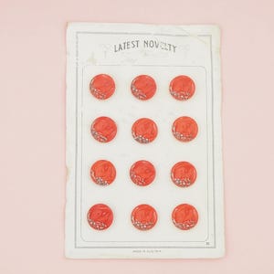 May include: A vintage sheet of red buttons with a decorative design, arranged in three rows of four. The sheet is labelled "LATEST NOVELTY" and "MADE IN AUSTRIA" on a pink background.