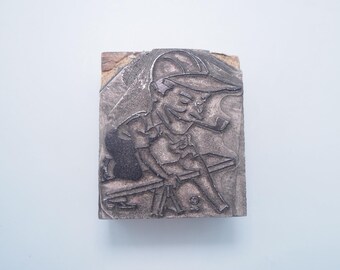May include: A vintage wood printing block with a metal relief of a cartoon character wearing a hat and sitting on a bench. The character is holding a pipe in their hand.
