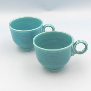 Può includere: Due tazze da tè in ceramica turchese con manici rotondi. Le tazze hanno un design classico con base a piede e dettagli orizzontali a coste attorno al corpo. Le tazze sono su uno sfondo bianco.