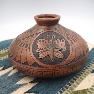 Può includere: Un vaso di ceramica marrone fatto a mano, con un corpo rotondo e una piccola apertura. Il vaso presenta un disegno a farfalla intagliato al centro, circondato da motivi geometrici. Il vaso è esposto su un tessuto a motivi blu e bianchi.