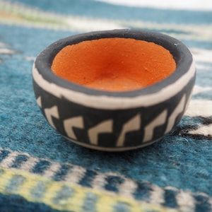 Può includere: Una piccola ciotola rotonda in ceramica con esterno nero, dettagli bianchi e interno arancione. La ciotola presenta un motivo geometrico attorno al bordo. La ciotola è appoggiata su un tessuto a fantasia blu e bianco.