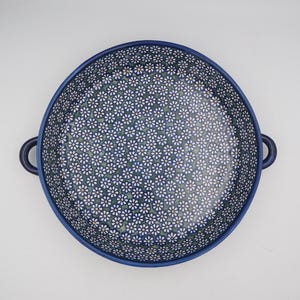 Puede incluir: Una fuente de horno redonda de cerámica con dos asas laterales, que presenta un patrón denso de pequeñas flores de margarita blancas con centros amarillos sobre un fondo azul oscuro y verde, vista desde arriba sobre un fondo blanco.