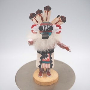 May include: A hand-carved Native American Kachina doll. The doll features a detailed painted design with a headdress, a white fur collar, and a patterned skirt. It stands on a small wooden base, showcasing traditional craftsmanship.