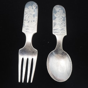 May include: A silver fork and spoon set with Mickey Mouse and Minnie Mouse engraved on the handles. The fork has four tines and the spoon has a rounded bowl.