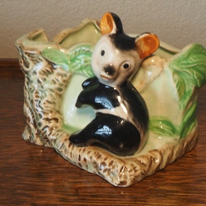Puede incluir: Un macetero de cerámica con forma de tronco de árbol con un koala negro y blanco sentado encima. El koala tiene orejas naranjas y está pintado en estilo de dibujos animados. El macetero es verde y marrón.