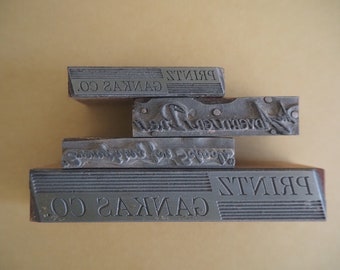 May include: A stack of four vintage printing blocks with the text "GAKAS CO. PRINTZ" on the bottom block and "PRINTZ" on the top block.