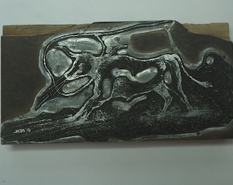 May include: A close-up of a woodblock print with a silver-colored design. The design is abstract and features a series of swirling lines and shapes. The woodblock is signed "C. WEAL" in the lower left corner.