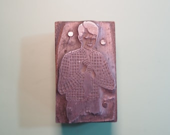 May include: A vintage printing block featuring a raised image of a person wearing a plaid shirt. The block is made of metal and has a brown patina.