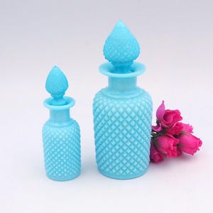May include: Two light blue glass perfume bottles with diamond-patterned exteriors and matching stoppers. A small bouquet of pink roses sits beside the bottles. The bottles are different sizes, with a textured surface.