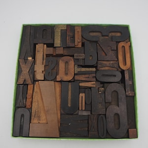 May include: A square collection of vintage wooden letterpress blocks arranged tightly within a green-rimmed box. The assortment features various letters, numbers, and symbols in different sizes, wood grains, and dark brown to light tan tones.