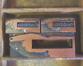 Op de afbeelding: Drie vintage drukblokken met koperen en metalen details. Twee blokken tonen het woord "establish" in omgekeerde volgorde, terwijl het derde blok een hand heeft die wijst. De blokken zijn gerangschikt in een houten bak, met een rustieke uitstraling.