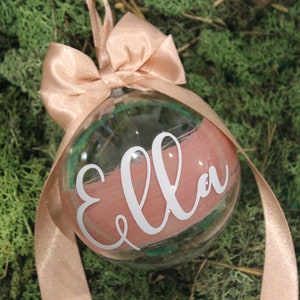 May include: A clear glass bauble with a pink and green paint design. The name "Ella" is written in white lettering on the bauble. The bauble is tied with a pink ribbon.