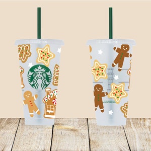 May include: Two clear plastic tumblers with green straws and a Christmas cookie design. The tumblers feature gingerbread men, star-shaped cookies, and a gingerbread house. One tumbler has the Starbucks logo.