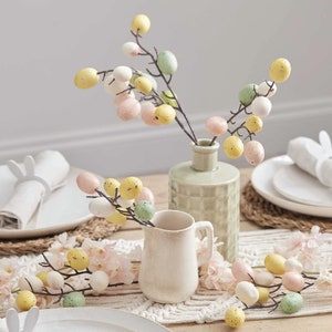 May include: A table setting with a white jug and a green vase filled with branches of pastel coloured Easter eggs. The branches are arranged on a white crocheted table runner.