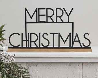 Può includere: Segno in metallo nero con le parole "Merry Christmas" in uno stile di font moderno. Il segno è seduto su una base di legno.