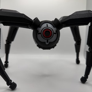 May include: A black, mechanical spider-like robot with articulated legs and a central body. The body features a gray and black design with a red circular detail, resembling a target or camera lens. The robot is set against a plain white background.