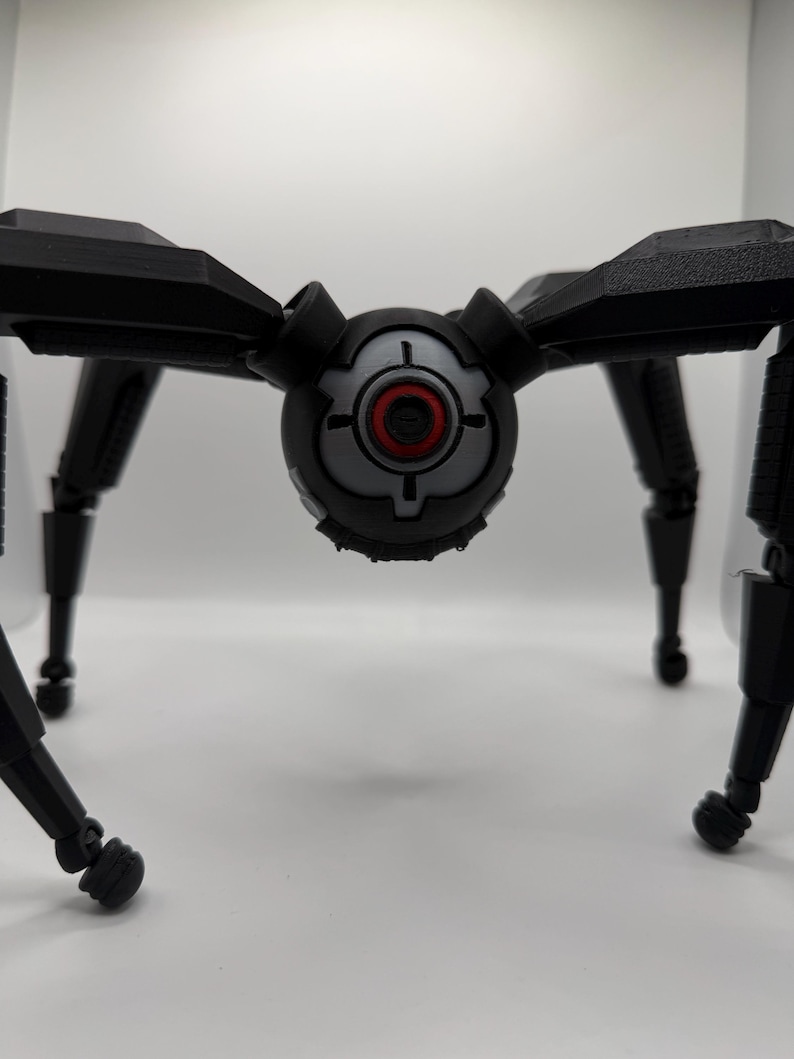 May include: A black, mechanical spider-like robot with articulated legs and a central body. The body features a gray and black design with a red circular detail, resembling a target or camera lens. The robot is set against a plain white background.