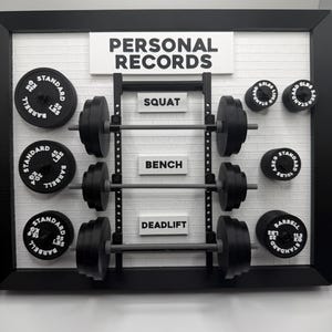 May include: A framed wall decor piece with the words "PERSONAL RECORDS" at the top. It features a weightlifting theme with barbells and weight plates labeled with weights in pounds. The words "SQUAT", "BENCH", and "DEADLIFT" are also displayed.
