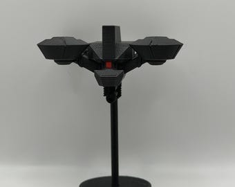 May include: A black, futuristic-looking model, possibly a drone or spacecraft, mounted on a black stand. The model has a central body with four wing-like structures and a small red accent. The stand has a circular base.
