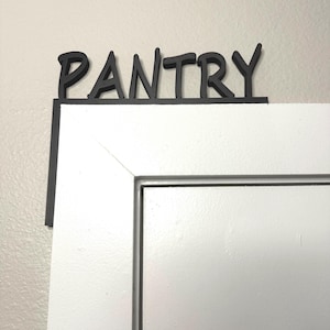 May include: Black wooden "PANTRY" sign mounted above a white door. The sign is in a bold, sans-serif font and is attached to a black rectangular frame. The door is white with a simple, modern design.