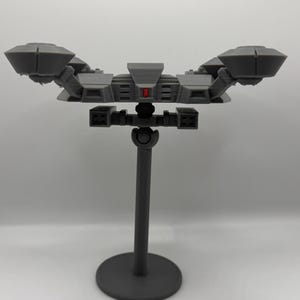 May include: A gray, 3D-printed model of a spacecraft, featuring a central body with angular details and two large, wing-like structures on either side. The model is mounted on a gray stand with a circular base. A small red accent is visible.