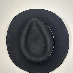 Puede incluir: Un sombrero fedora negro con ala ancha. El sombrero tiene una copa redondeada y una banda de color beige en contraste alrededor de la base de la copa. El sombrero está hecho de un material suave.