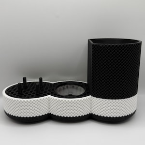 May include: A black and white 3D-printed desk organizer. The organizer has a cylindrical cup holder, a section with three small posts, and a circular opening. The base is white with a textured pattern, and the top sections are black.