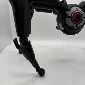 May include: A black, articulated robot toy with a spherical head featuring a red and gray eye-like design. The robot has multiple jointed limbs and a three-legged stance, suggesting a mechanical or futuristic theme.