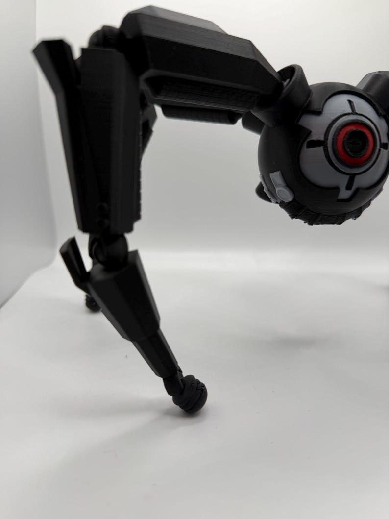 May include: A black, articulated robot toy with a spherical head featuring a red and gray eye-like design. The robot has multiple jointed limbs and a three-legged stance, suggesting a mechanical or futuristic theme.