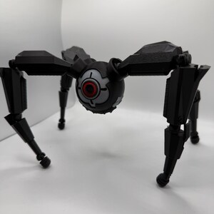 May include: A black robotic spider toy with a central spherical body and eight articulated legs. The central body features a red and gray eye-like design. The legs are segmented and end in rounded feet.