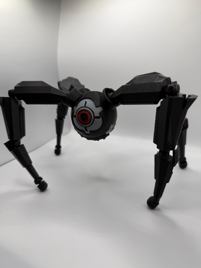 May include: A black robotic spider toy with a central spherical body and eight articulated legs. The central body features a red and gray eye-like design. The legs are segmented and end in rounded feet.